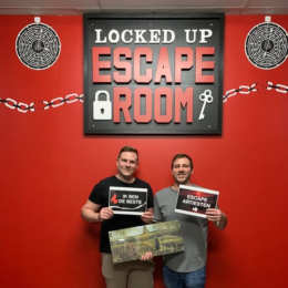 Twee mannen houden borden en een schilderij vast voor het Locked Up Escape Room-bord op een rode muur, samenwerking met Belimi