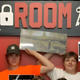 Groep jongeren in oranje shirts houdt borden vast met de tekst King, Team, Net op tijd en MVP, voor een rode escape room-muur